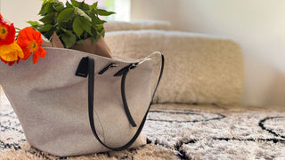 Beige tote bag with black straps on a textured surface with flowers and a plant in the background.