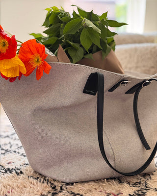 Gray tote bag with black handles and a plant inside, placed on a patterned surface with flowers in the foreground.