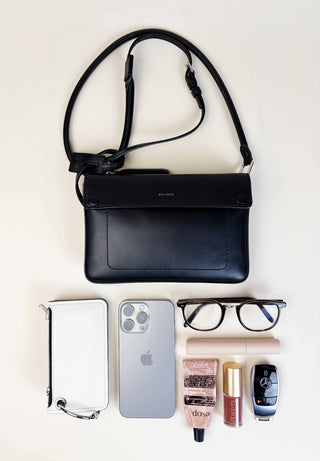 Black leather crossbody bag with phone, wallet, glasses, and beauty products on a white background